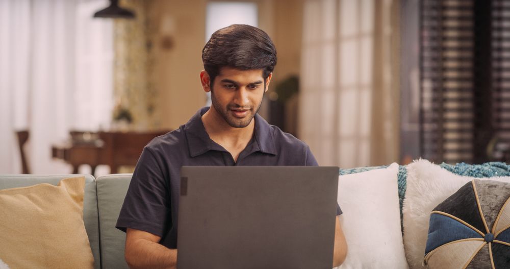 Young Indian Man Focused on Remote Work, Using Laptop on Sofa at Home.