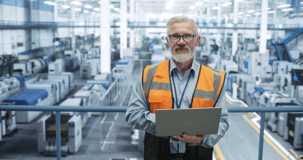 Portrait of a Technician with a Beard and Glasses Using Laptop Computer and Looking at Camera. Industrial Male Specialist Wearing a Protective Vest, Working in a Modern Electronics Factory