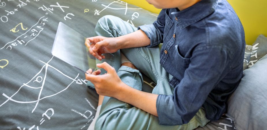 Unrecognizable young kid holding digital display with glass screen sitting over his bed