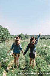 Two women wearing hats and holding hands and walking on grass field 0KLa75