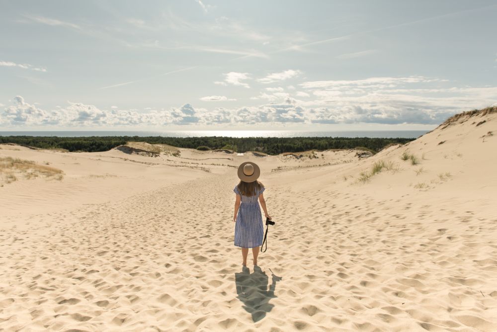 Back view of adult woman in straw hat and dress with camera walking on sand dune