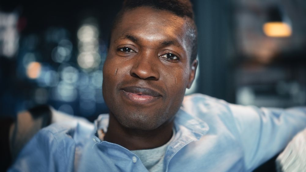 Close-up Face Portrait of the Young Black Man Smiling and Laughing at Something Funny on TV while Sitting on a Sofa in the Evening.