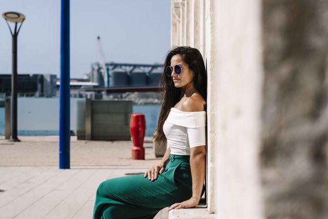 Side view of a young hispanic woman with sunglasses sitting on a stone bench against wall looking away