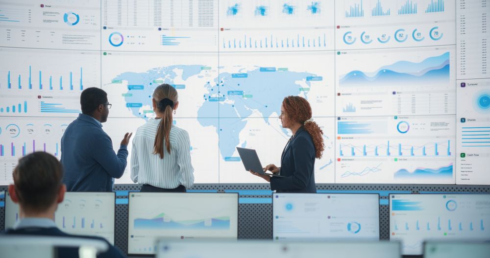 Diverse Team Of Specialists From Analytics Department Discussing Global Distribution In Front of Big Digital Screen With World Map In Monitoring Office.