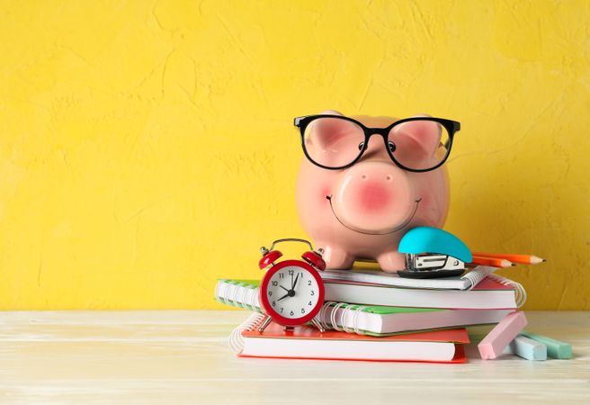 Happy piggy bank with glasses, and school accessories on wooden table against color background, space for text. Finance, saving money
