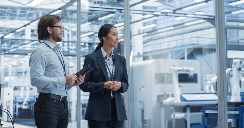 Two Diverse Engineers Walking in Office, Using Tablet Computer at an Electronics Factory. Female and Male Specialists Chatting, Monitoring Robots on Modern Assembly Line.