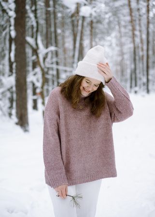 Woman in pink sweater and light cap smiling and standing in snow covered wood