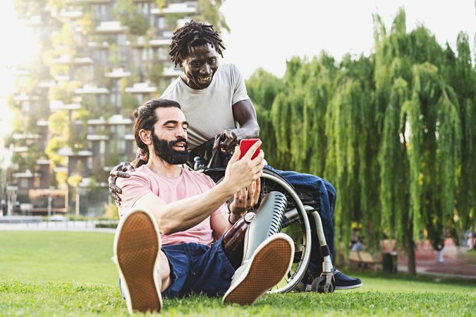 Two friends, one Hispanic with a prosthetic leg and one Black in a wheelchair are relaxing outdoors chatting together and using smartphones