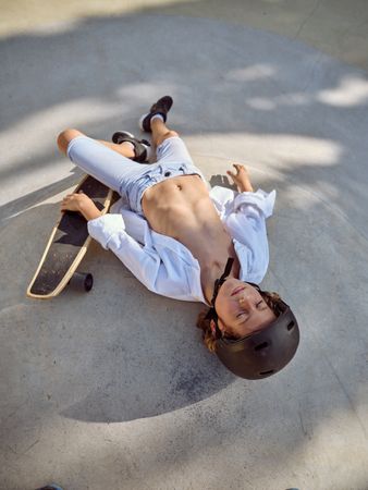 Young boy resting on asphalt with skateboard in hand while helme