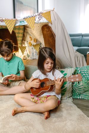 Little girl playing ukulele while boy reading book on handmade teepee in living room at home