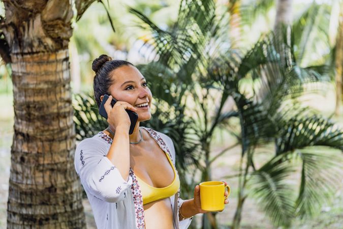 Cheerful woman talking on phone while drinking coffee by palm tree