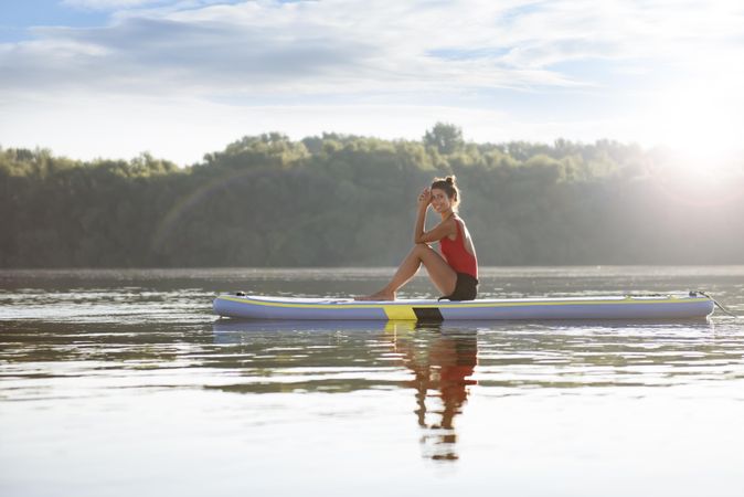 Happy woman sitting relaxing on paddleboard near lake in the morning
