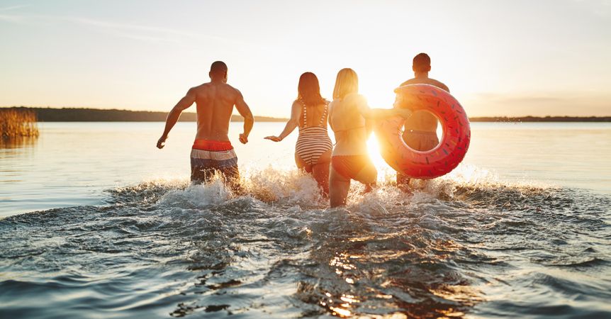 Multi-ethnic group of friends running into the lake at sunset with floating ring