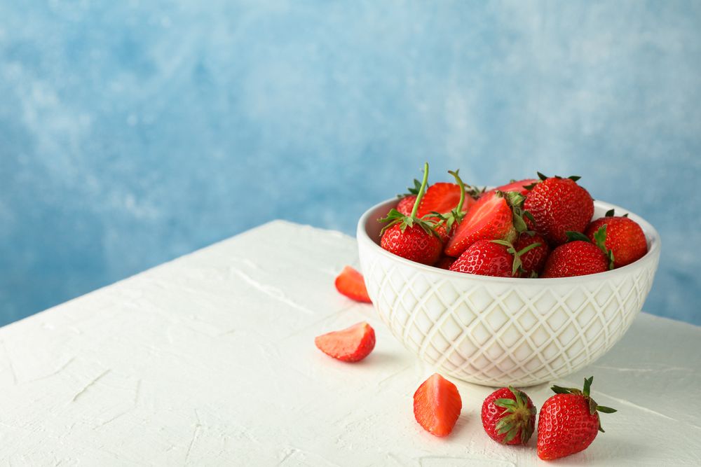 Bowl with fresh strawberries on table against blue background, space for text. Summer sweet fruits and berries