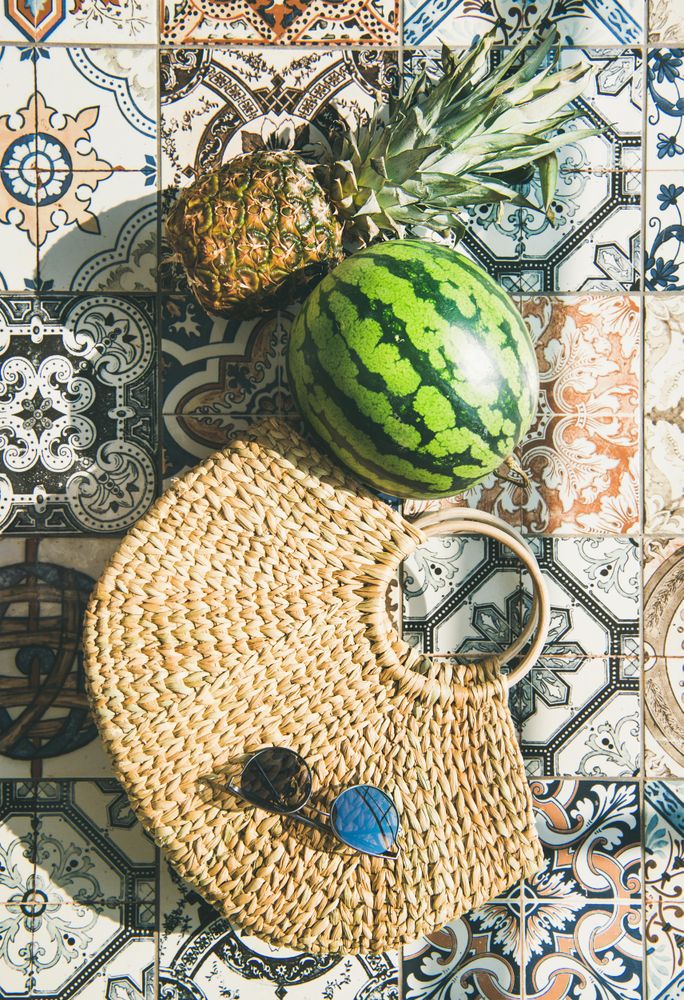 Thatched bag on pattered tiles with sunglasses, watermelon and pineapple, with shadows