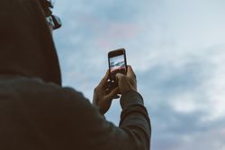 Rearshot of man on cliff taking photo on phone at sunset
