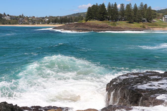 Waves crash against rocky shore with lush green hills and sandy beach in the background under clear blue sky