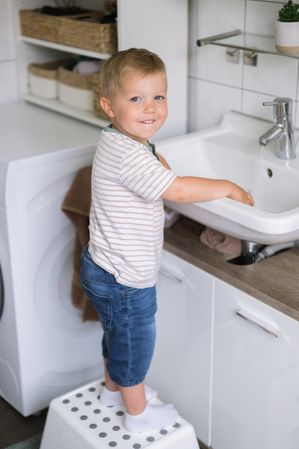 Cute child boy standing on stool near washing sink in daylight