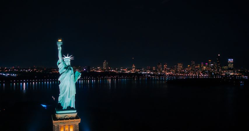 Aerial Fly-By Around the Statue of Liberty. Helicopter Photo of the Patriotic Monument in Beautiful Night Setting. Ferry Boats with Lights Transporting Tourists To and From Manhattan