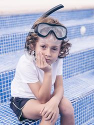 Adorable preteen boy in snorkeling mask sitting on tiled poolsid