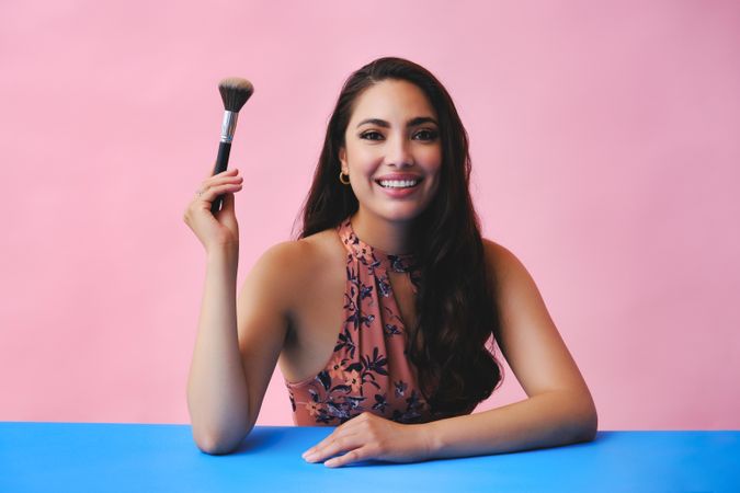 Hispanic woman with long brown hair holding large make up brush up and smiling at camera