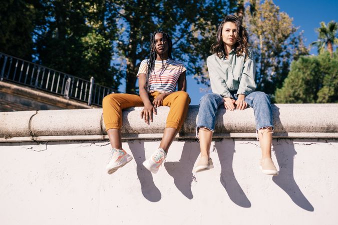 Serious female friends sitting on outdoor wall on nice day