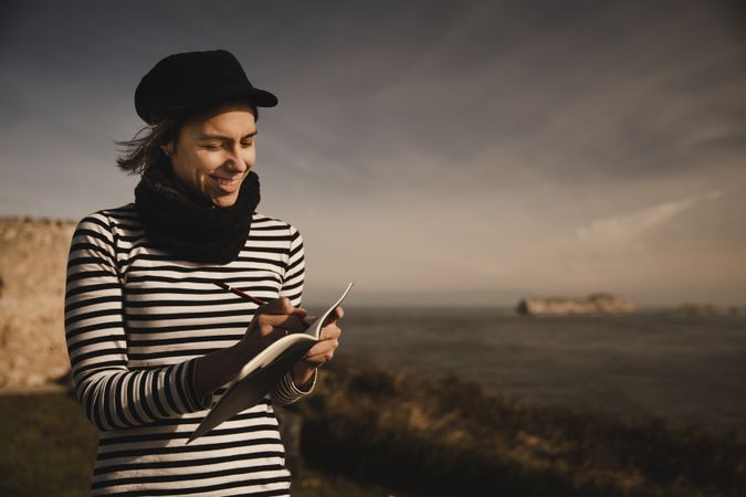 Woman writing in notebook on bench on shore near water