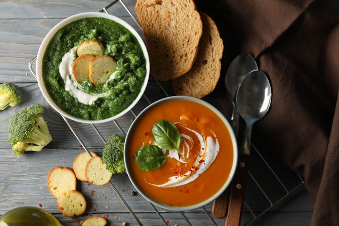 Two bowls of orange and green soup on wooden table with crackers, vegetables and spoons