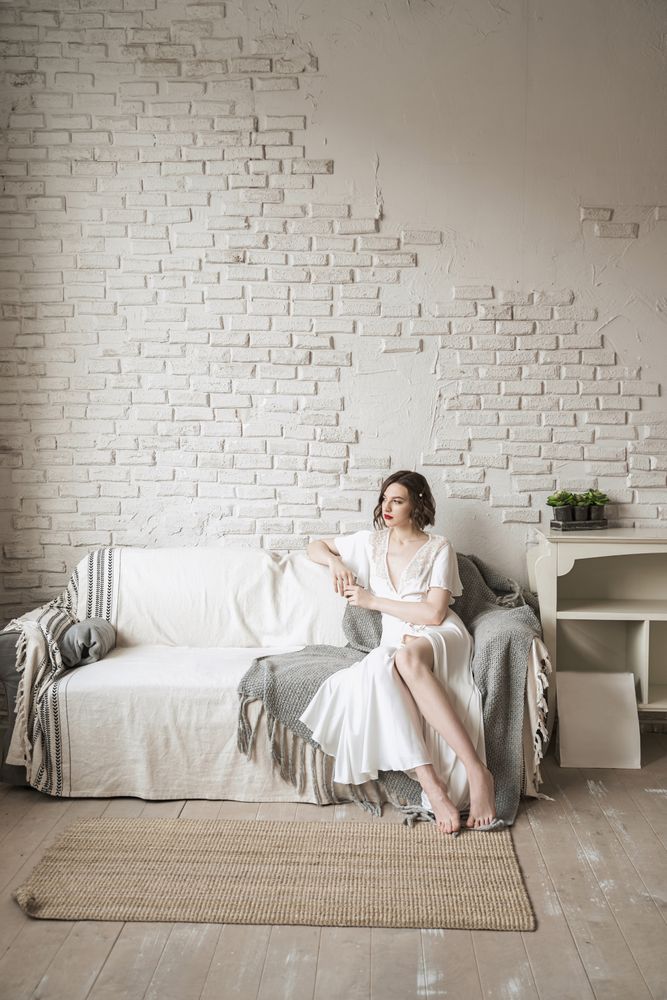 Pensive adult woman looking away and dreaming while sitting on sofa and having hot drink in light modern living room