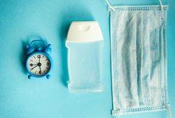Clock, hand sanitizer and facemask on blue pastel background