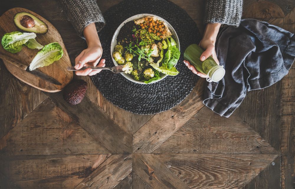 Healthy vegetarian bowl pictured on table with smoothie on side and hands with fork, copy space