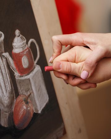 Hand of teacher guiding student as he draws a still life of tea pot and apple
