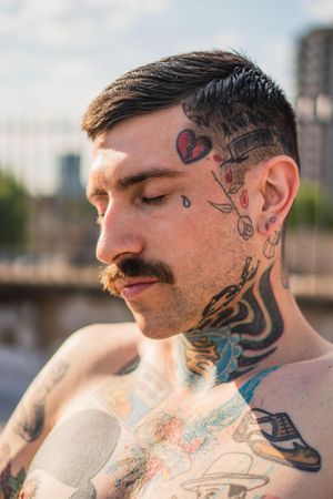 Shirtless white hipster man with mustache closing eyes in sunlight with face and neck tattoos