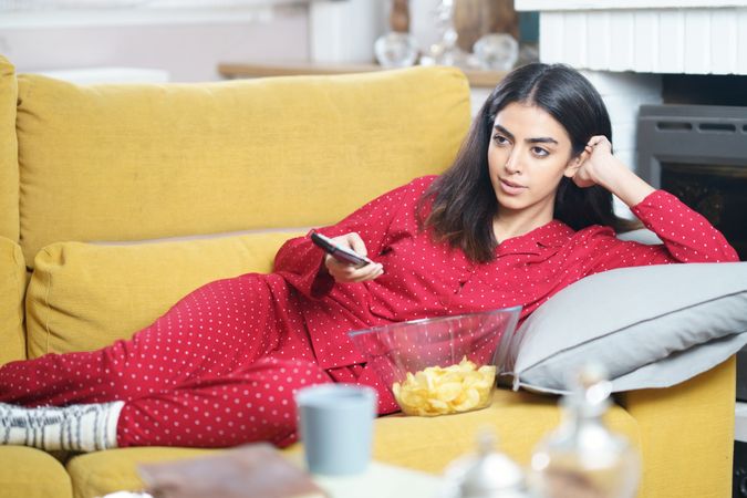 Woman in red pajamas relaxing on sofa with bowl of chips and remote control