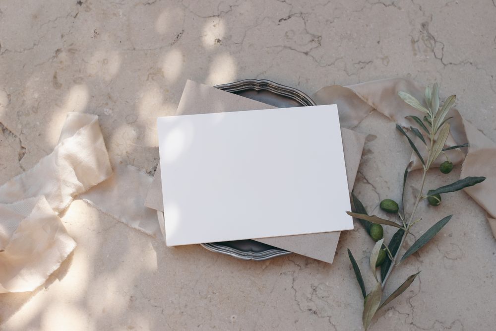 Blank greeting card, envelope on silver plate with olive tree branches on beige marble background