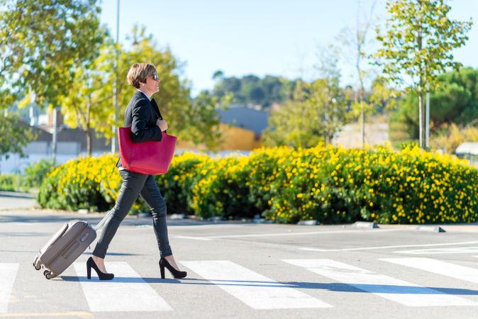 Woman in heels walking confidently across street with roller suitcase behind her