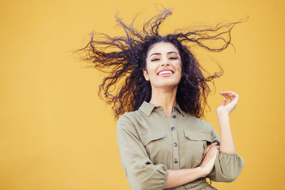 Female in army green jumpsuit smiling in front of mustard wall with arms folded & hair blowing back