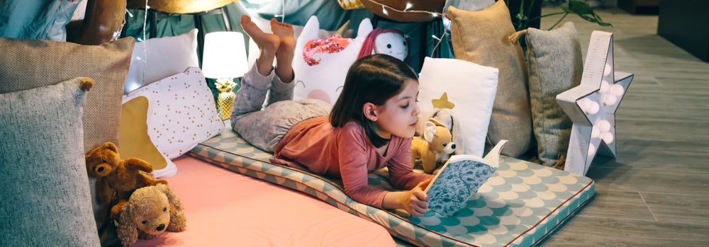 Banner of little girl reading and relaxing lying over sleeping mat in a cozy tent at home