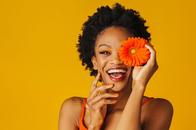 Joyful Black woman holding a flower over her eye with her hand to her chin