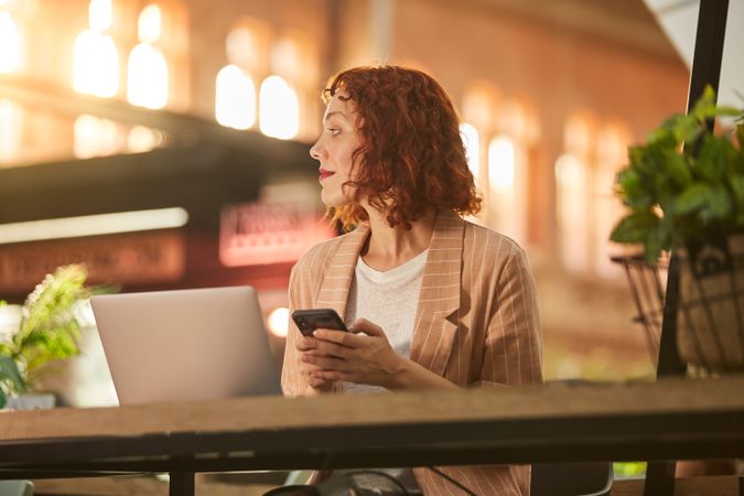 Cheerful young woman working on laptop in cafe and browsing phone