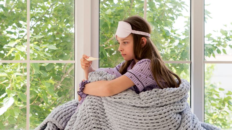 Girl sitting by window, looking pensive with electronic thermometer