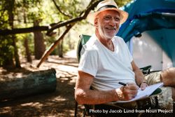 Smiling older man sitting in front of a tent and writing in a book 5aEOa5