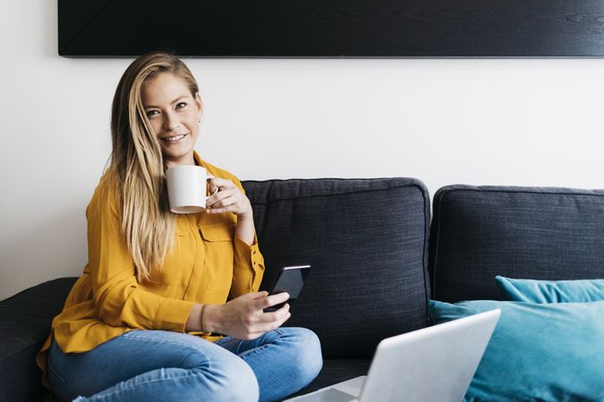 Beautiful adult female sitting on sofa holding smartphone on sofa with cup of coffee