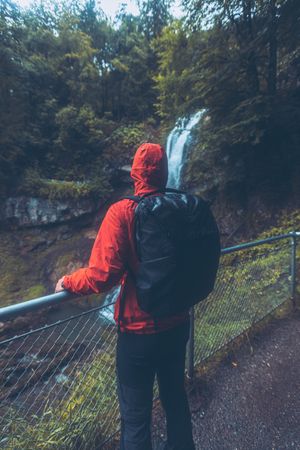 Back view of a man in red jacket with backpack standing against waterfall