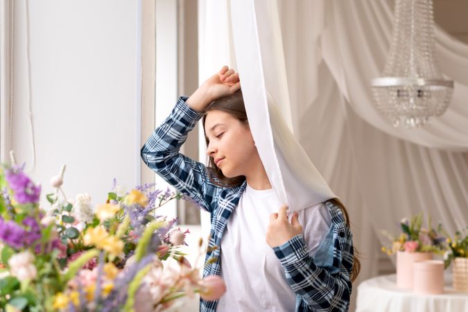 Girl gently holding white curtain near window at home indoors.