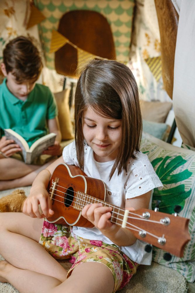 Portrait of smiling little girl playing ukulele while boy reading book on handmade teepee at home