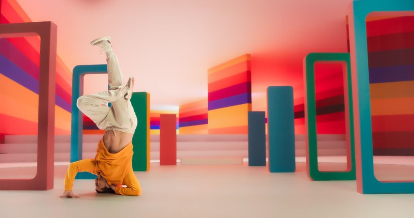 Male Dancer in Bright Sweater Performing Swift Breakdancing Moves With A Freeze in a Colorfully Decorated Studio. Professional B-Boy Showcasing Talent and Skills, Surrounded by Geometric Decoration