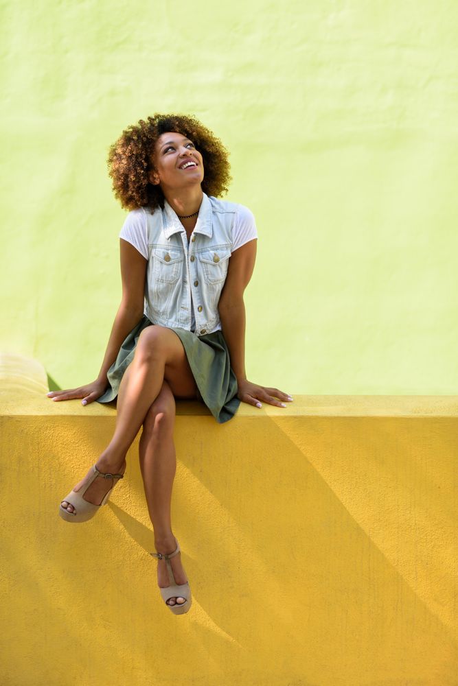 Woman in skirt and heels looking up while sitting on yellow green wall