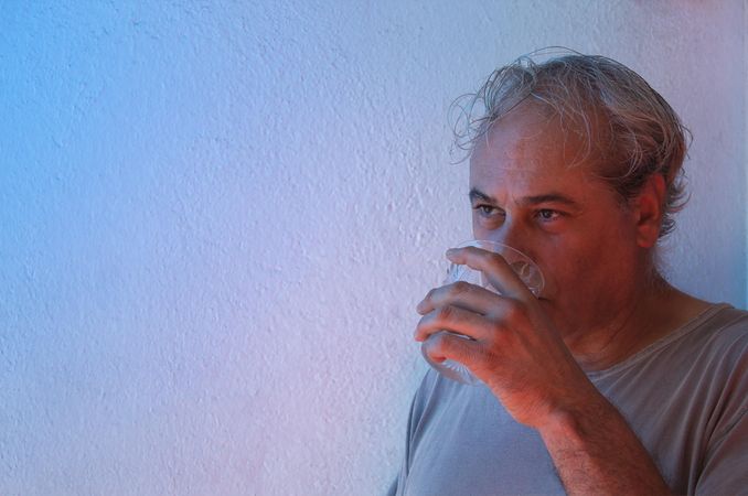 Portrait of middle aged man in gray shirt drinking water against light background in UV lit studio