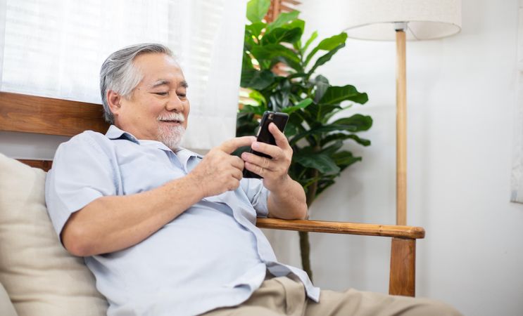 Happy older man smiling and sitting on couch using smartphone